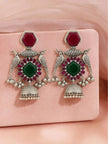 Model wearing and holding oxidised silver bird jhumka earrings with green and red stones against a neutral background