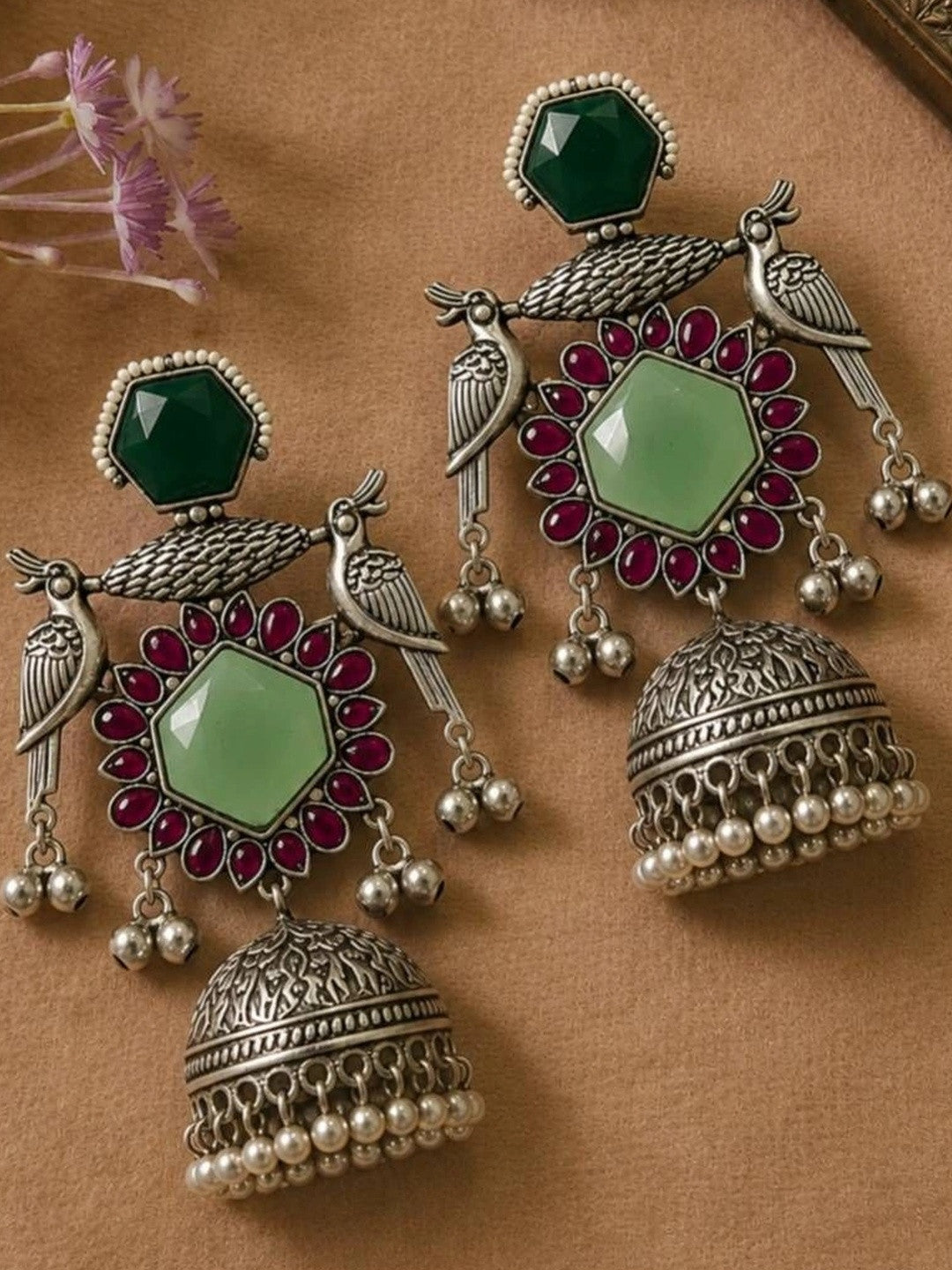 Pair of oxidised silver peacock jhumka earrings with green and ruby-red stones laid flat on a beige surface