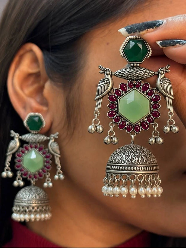 Woman holding one oxidised silver peacock jhumka earring close to her face showing full detail and scale