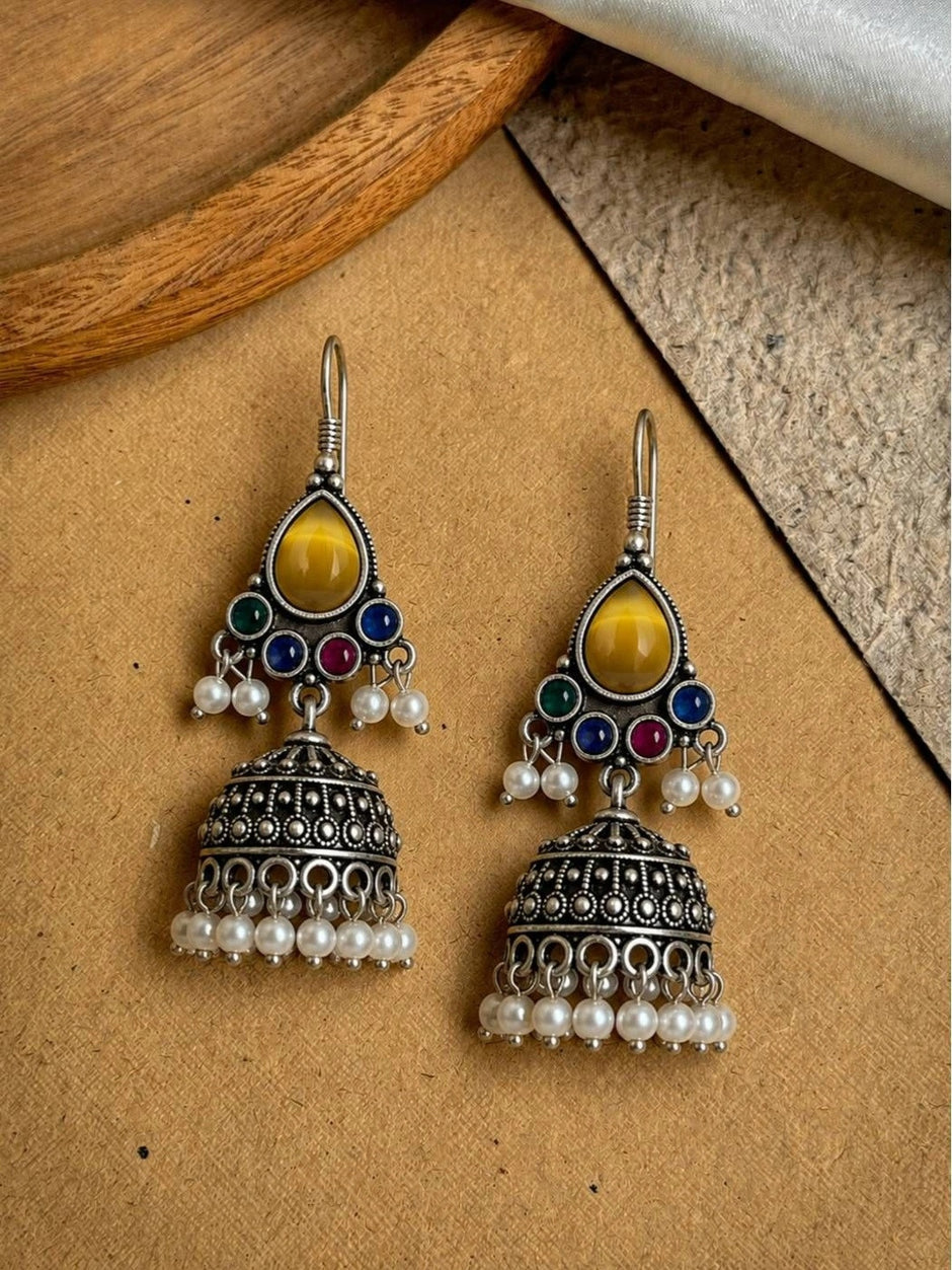Pair of oxidised silver jhumka earrings with yellow teardrop stone, multicolour cabochons, and white pearl drops on a beige surface