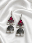 Pair of oxidised silver jhumka earrings with magenta teardrop stones and white pearl drops laid flat on ivory satin fabric