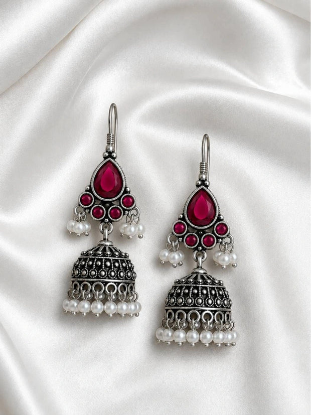 Pair of oxidised silver jhumka earrings with magenta teardrop stones and white pearl drops laid flat on ivory satin fabric