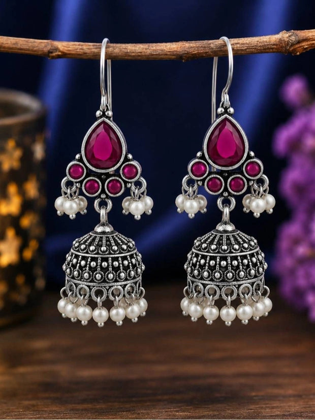 Both oxidised silver ruby stone jhumka earrings hanging from a wooden branch against a dark blue background