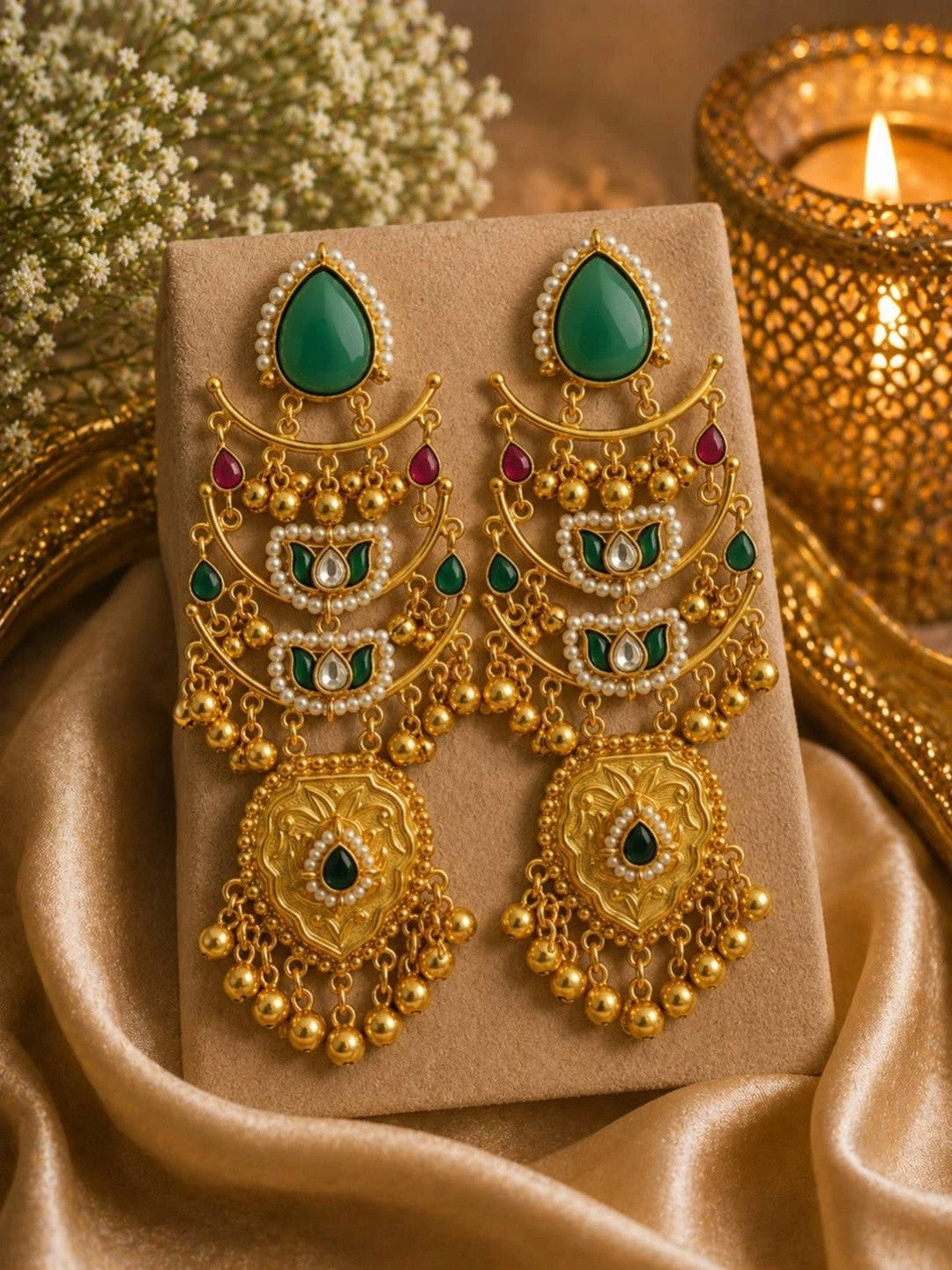 Pair of gold-plated kundan chandbali earrings with emerald green stones and ghungroo tassels displayed on a beige jewellery card