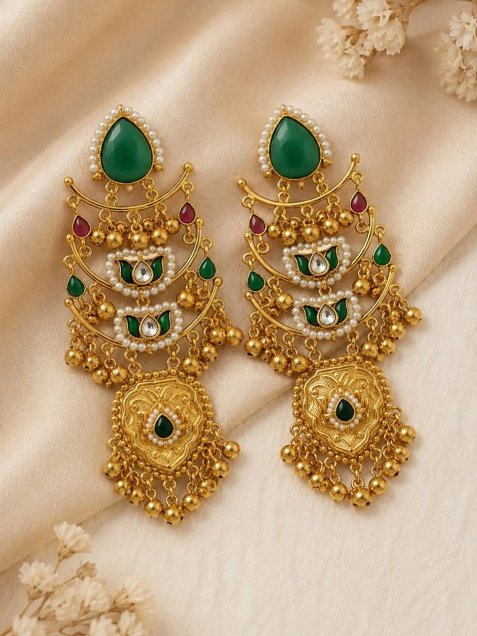 Two gold-plated chandbali earrings with green and red stones laid flat on cream silk fabric with dried flowers