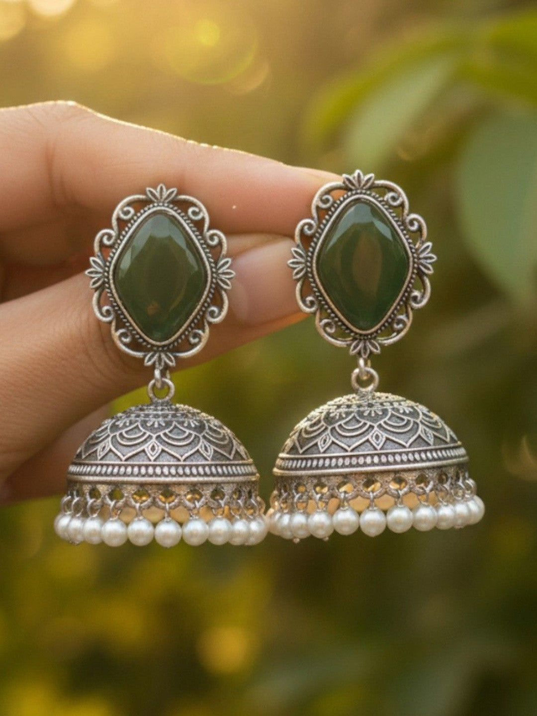 Hand holding a pair of oxidised silver jhumka earrings with green stone tops and white pearl fringe against bokeh green background