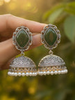Hand holding a pair of oxidised silver jhumka earrings with green stone tops and white pearl fringe against bokeh green background