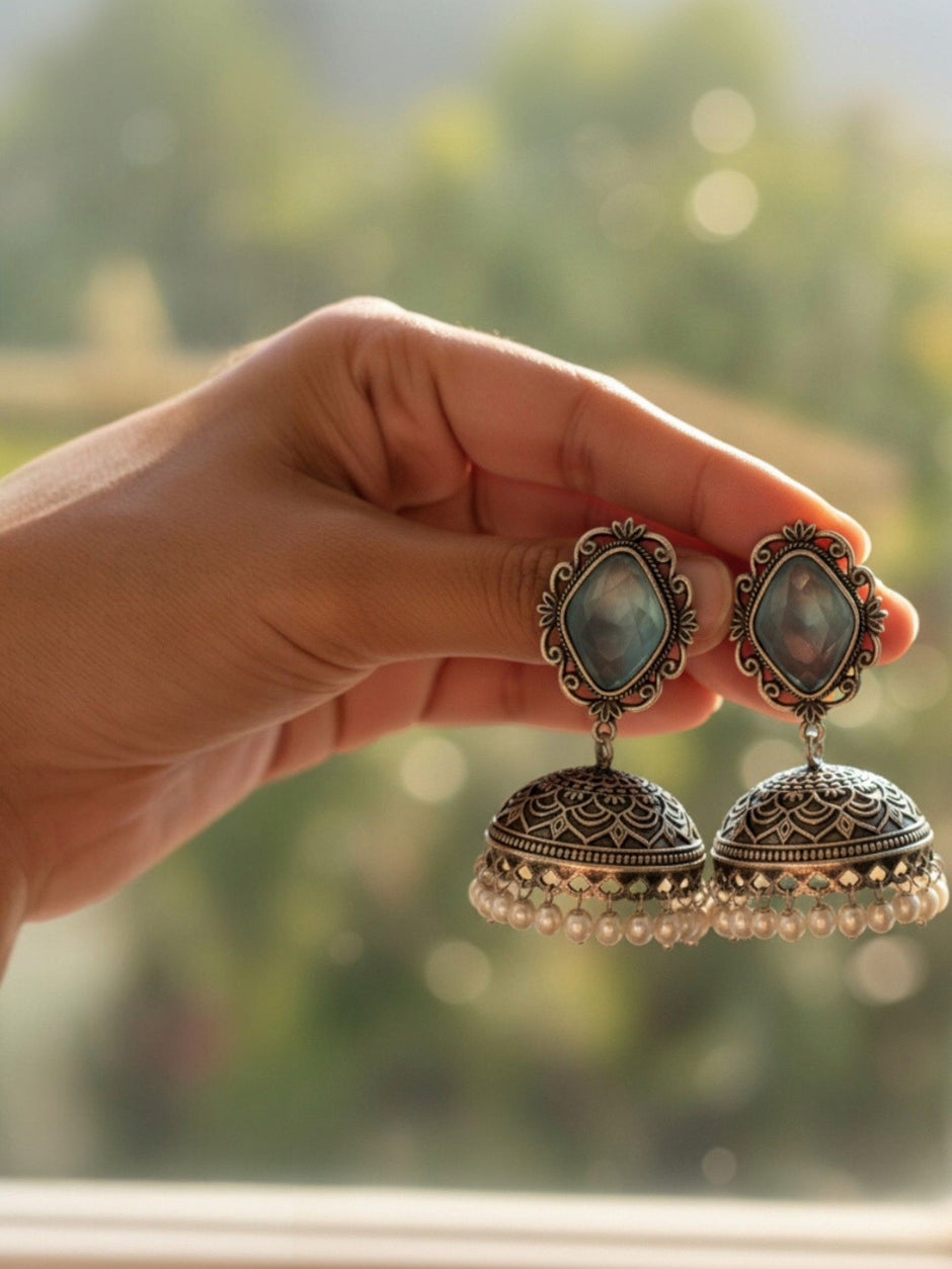 Hand holding a pair of oxidised silver jhumka earrings with blue stone tops and pearl drop fringe against a soft green bokeh background