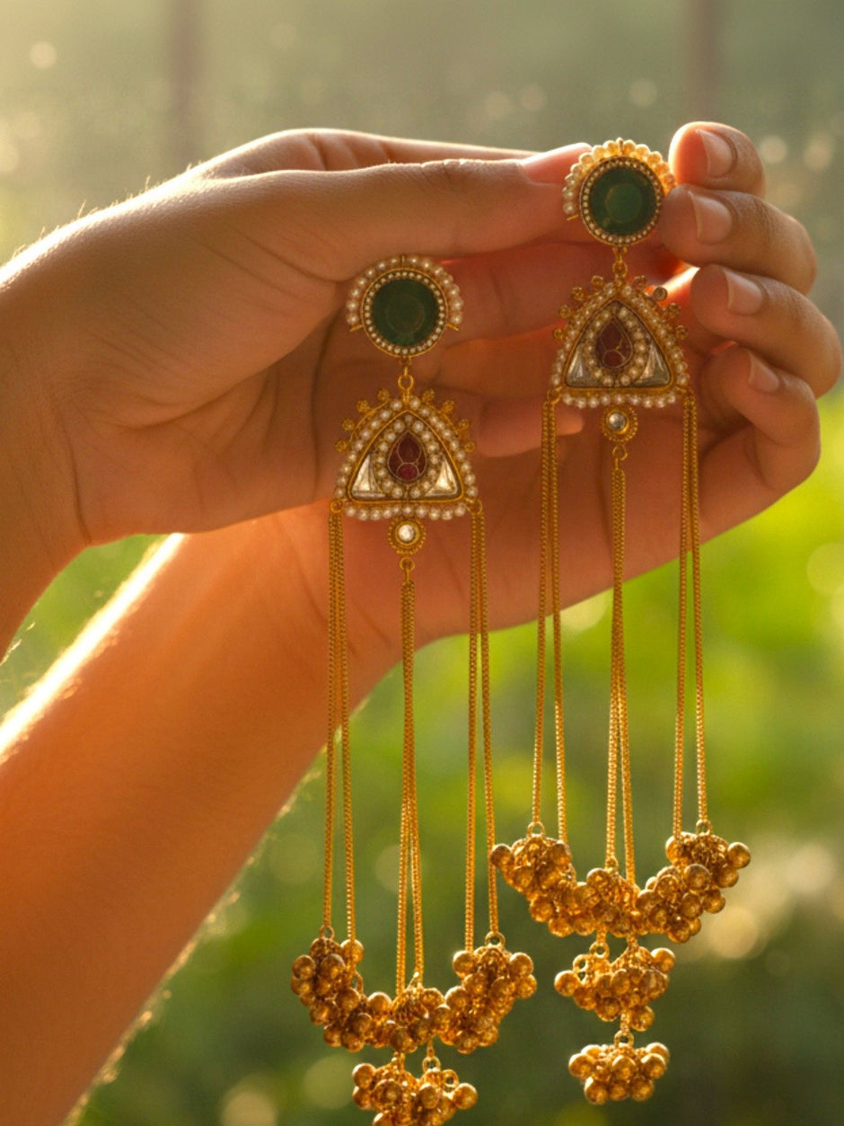 Hand holding two gold-plated kundan chandbali earrings with long chain and ghungroo clusters in warm sunlight