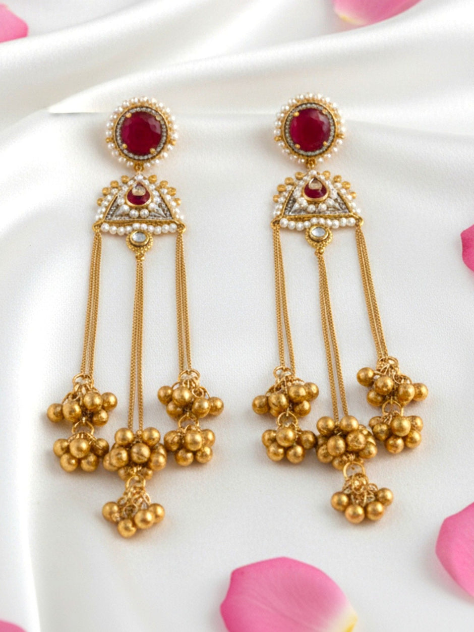 Pair of gold-plated kundan tassel earrings with ruby-red stone, pearl border, and clustered ghungroo drops on white satin