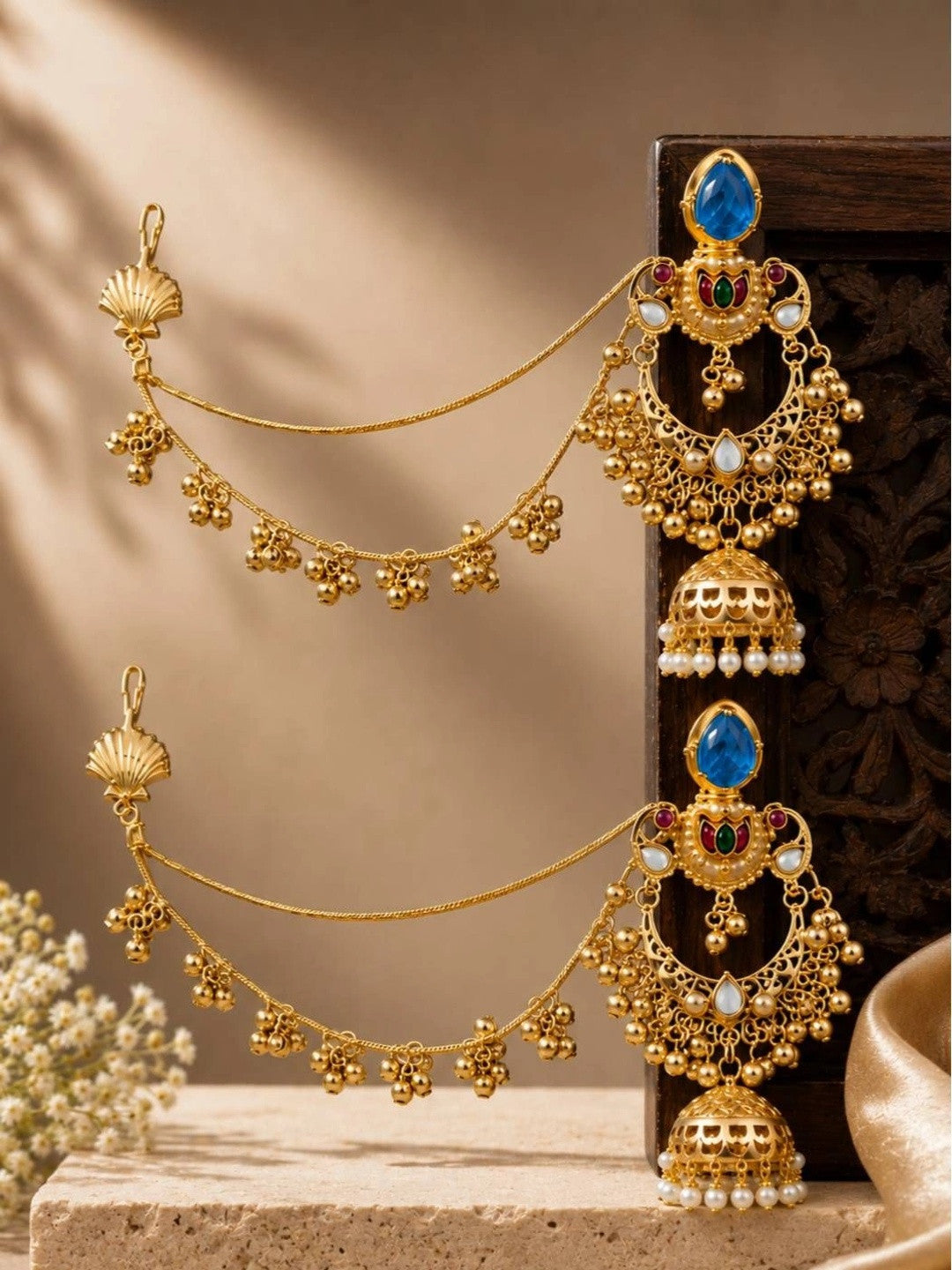 Pair of gold-plated chandbali jhumka earrings with blue stone tops and ghungroo hair chains displayed on a carved wooden block