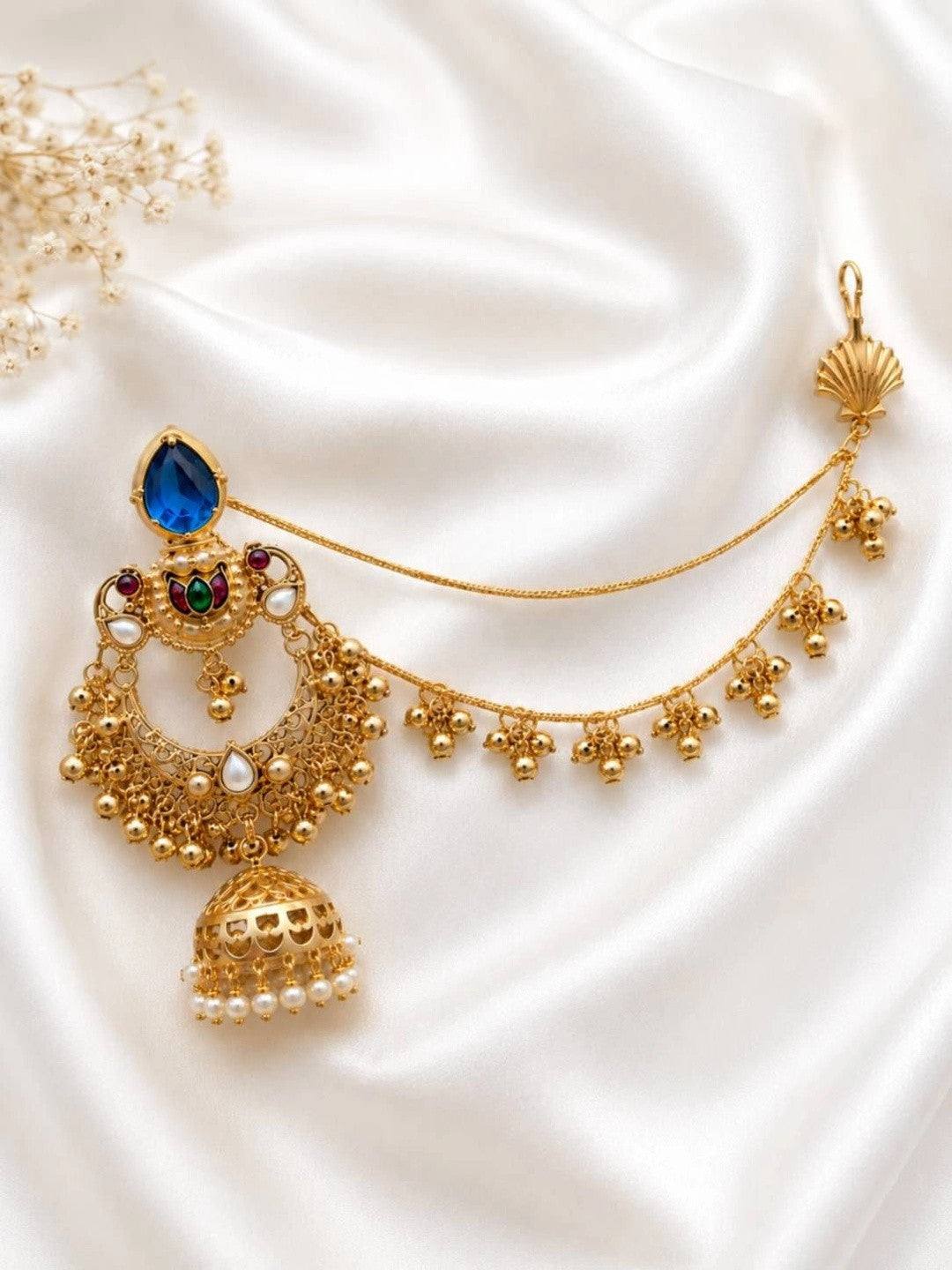 Single gold-plated chandbali jhumka earring with blue stone, ruby and emerald accents, and draped ghungroo hair chain on white satin