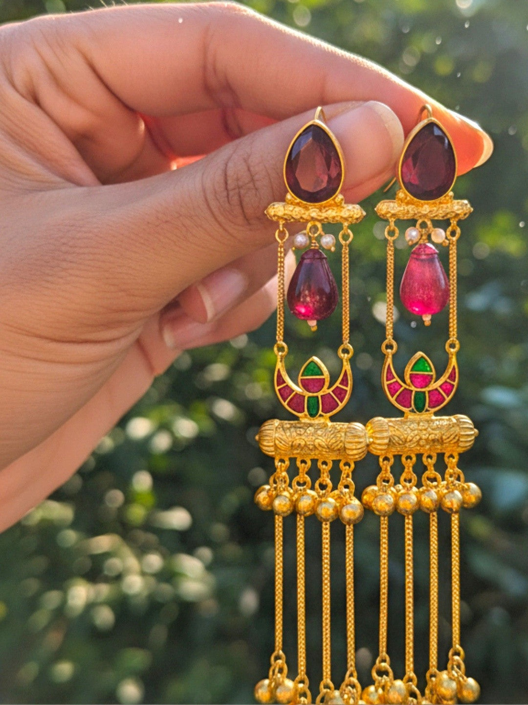 Close-up of gold tassel jhumkas with red and green meenakari crescent and ghungroo chains outdoors