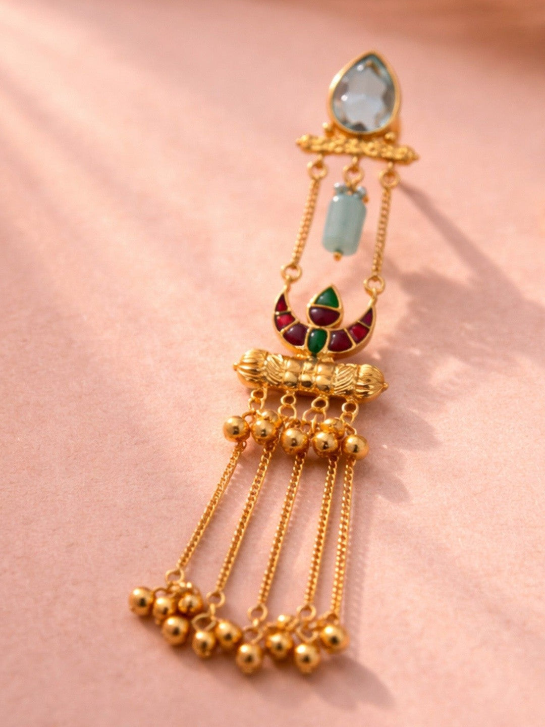 Single gold-plated meenakari chandelier earring laid flat on a pink surface showing blue teardrop stone and ghungroo tassels