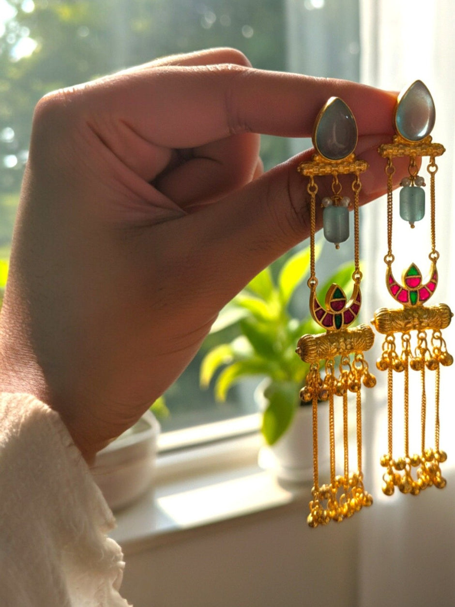 Hand holding pair of gold-plated meenakari tassel earrings near a sunlit window showing movement and length