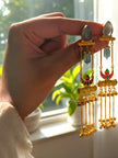 Hand holding pair of gold-plated meenakari tassel earrings near a sunlit window showing movement and length