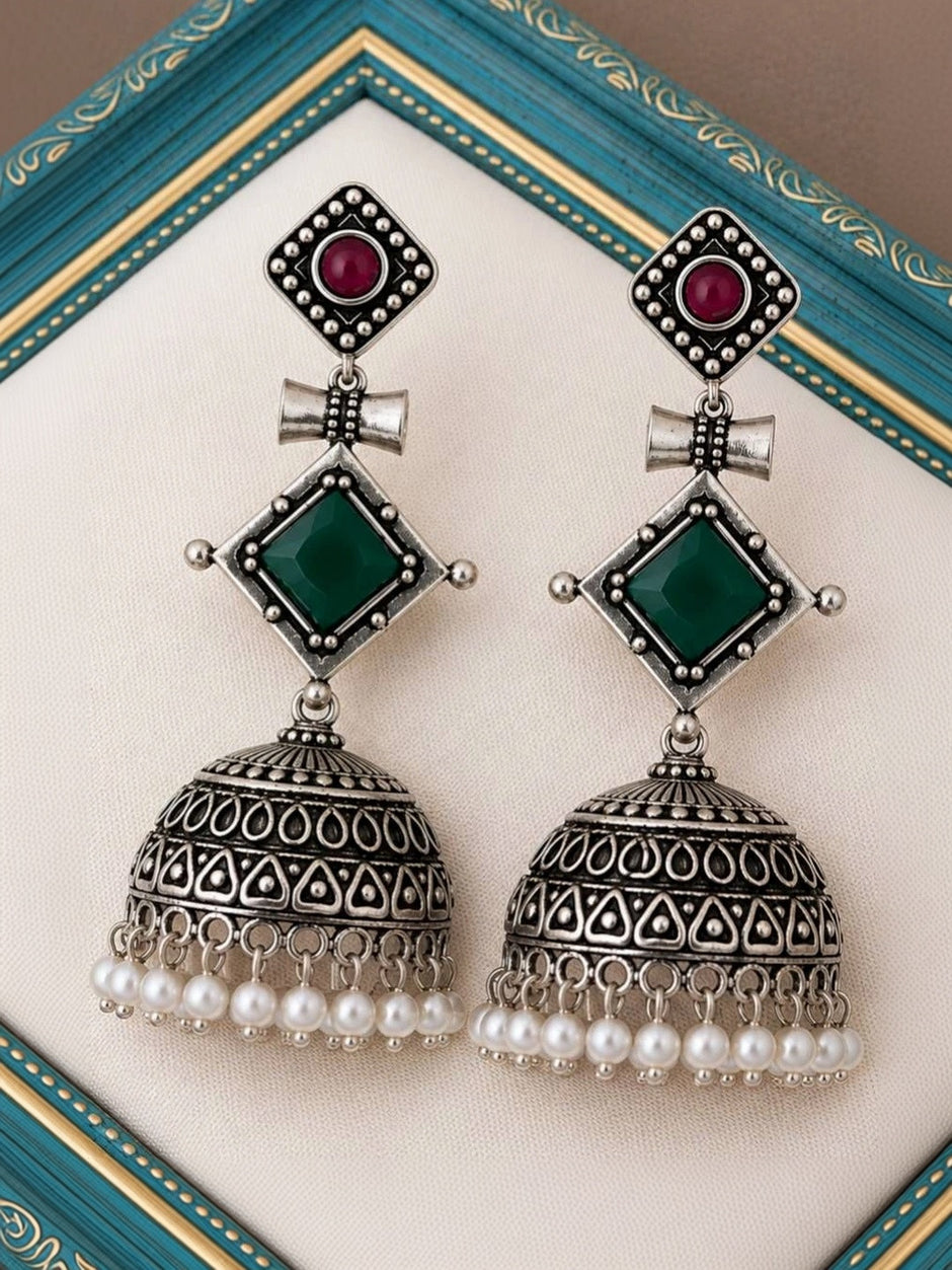 Pair of oxidised silver jhumka earrings with ruby-red and green stones, displayed on a teal decorative frame