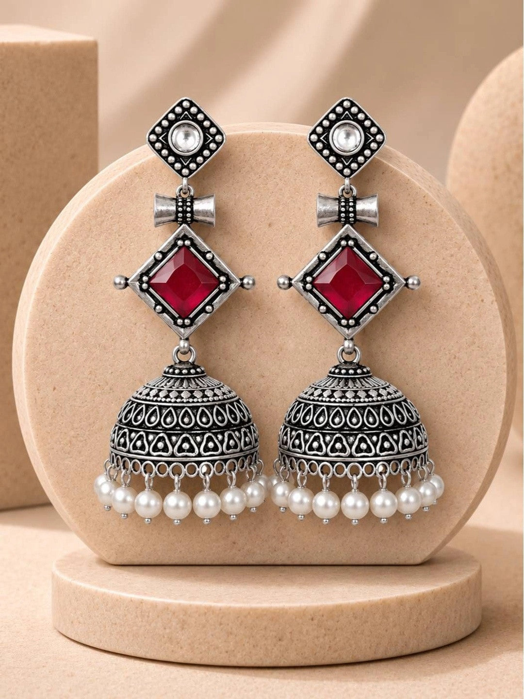 Pair of oxidised silver geometric jhumka earrings with red stone and pearl drops displayed on a beige disc stand
