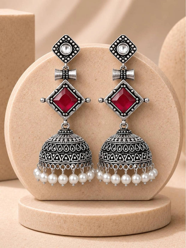 Pair of oxidised silver geometric jhumka earrings with red stone and pearl drops displayed on a beige disc stand