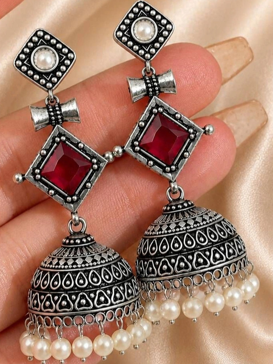 Close-up of two geometric jhumka earrings held in a hand showing engraved dome detail and red stone