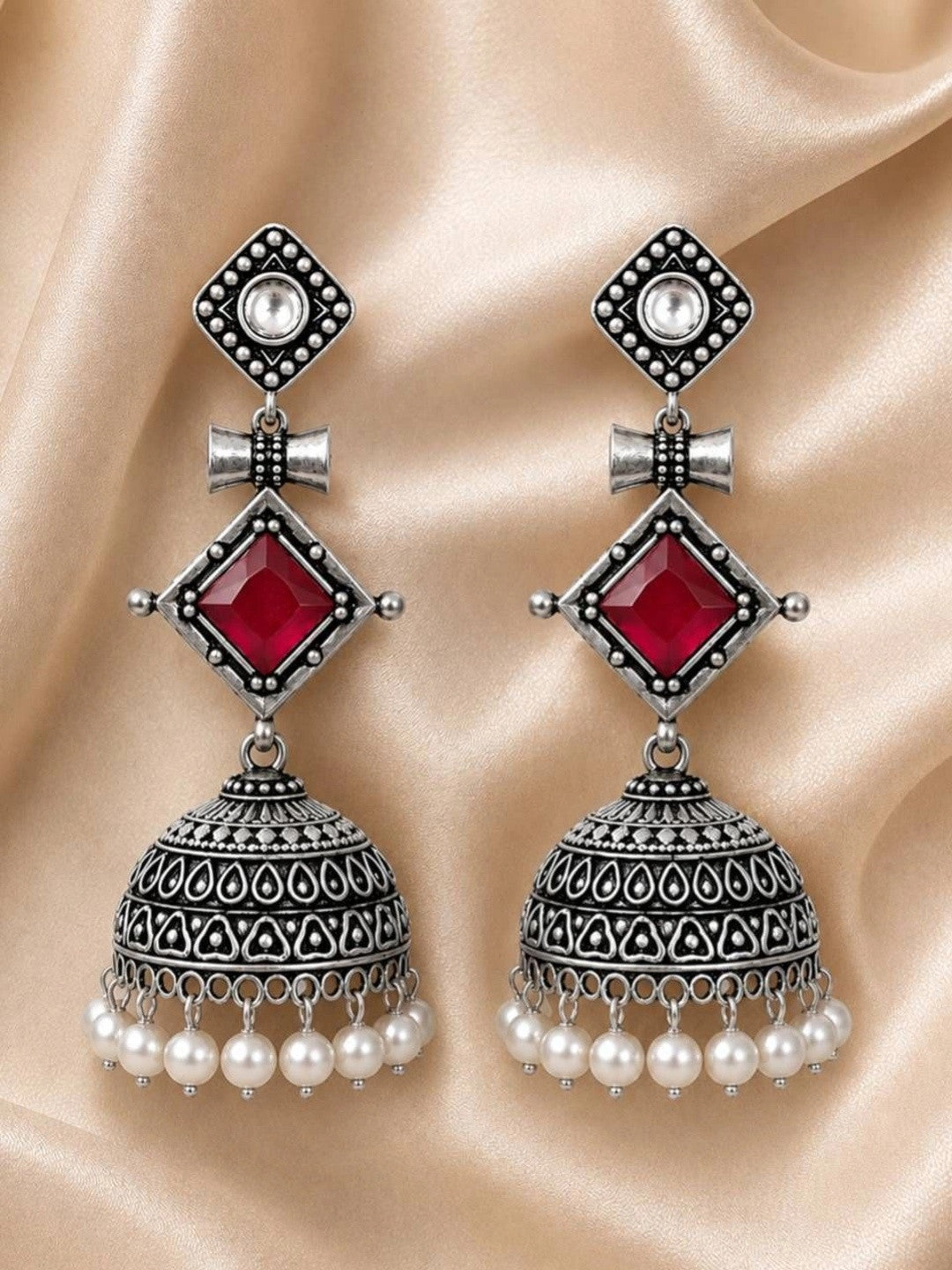 Pair of oxidised silver jhumka earrings with crimson stone and pearl drops laid flat on gold satin fabric