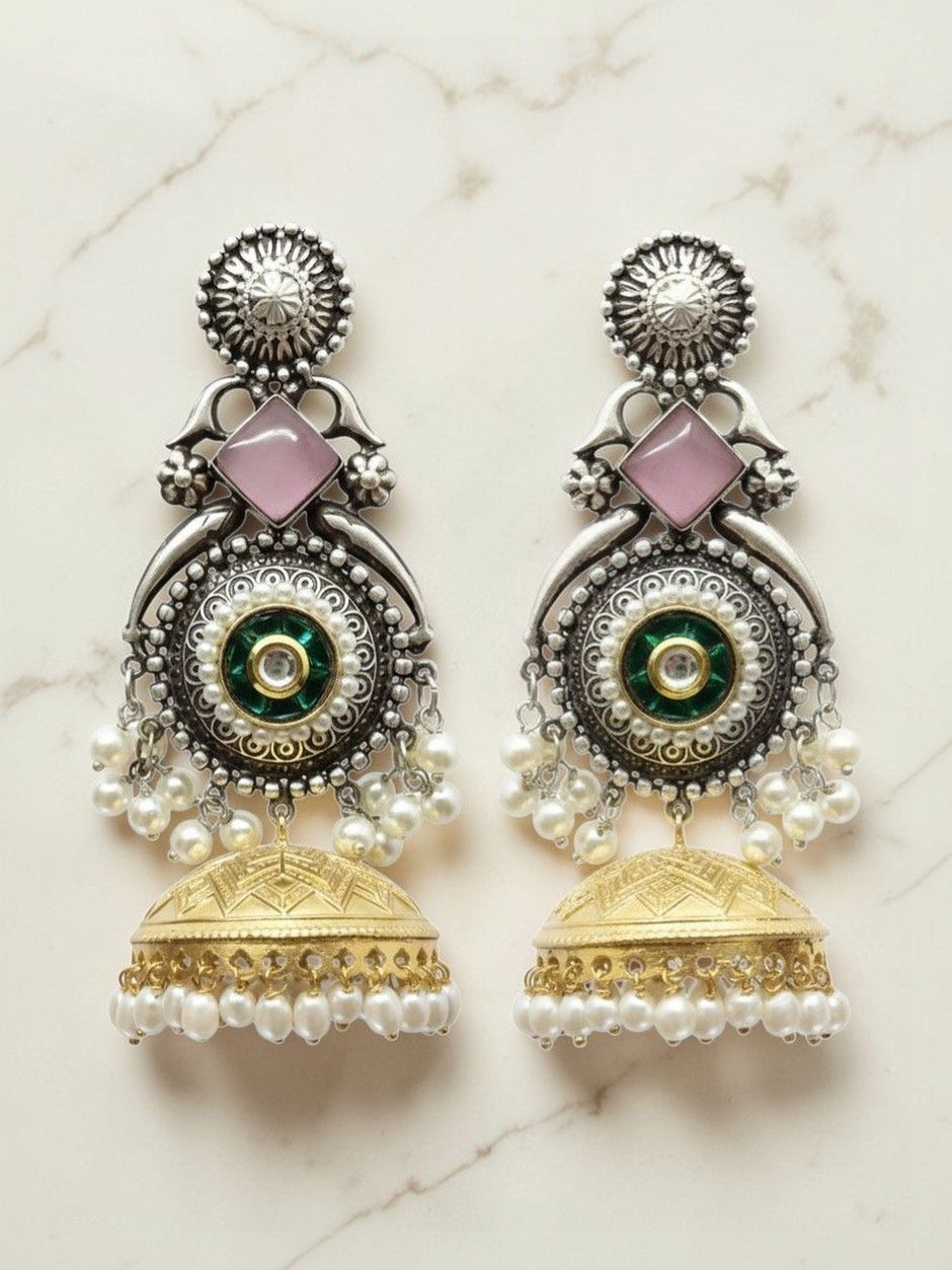Pair of oxidised silver kundan jhumka earrings with pink stone, pearl drops, and gold-tone bell on marble background