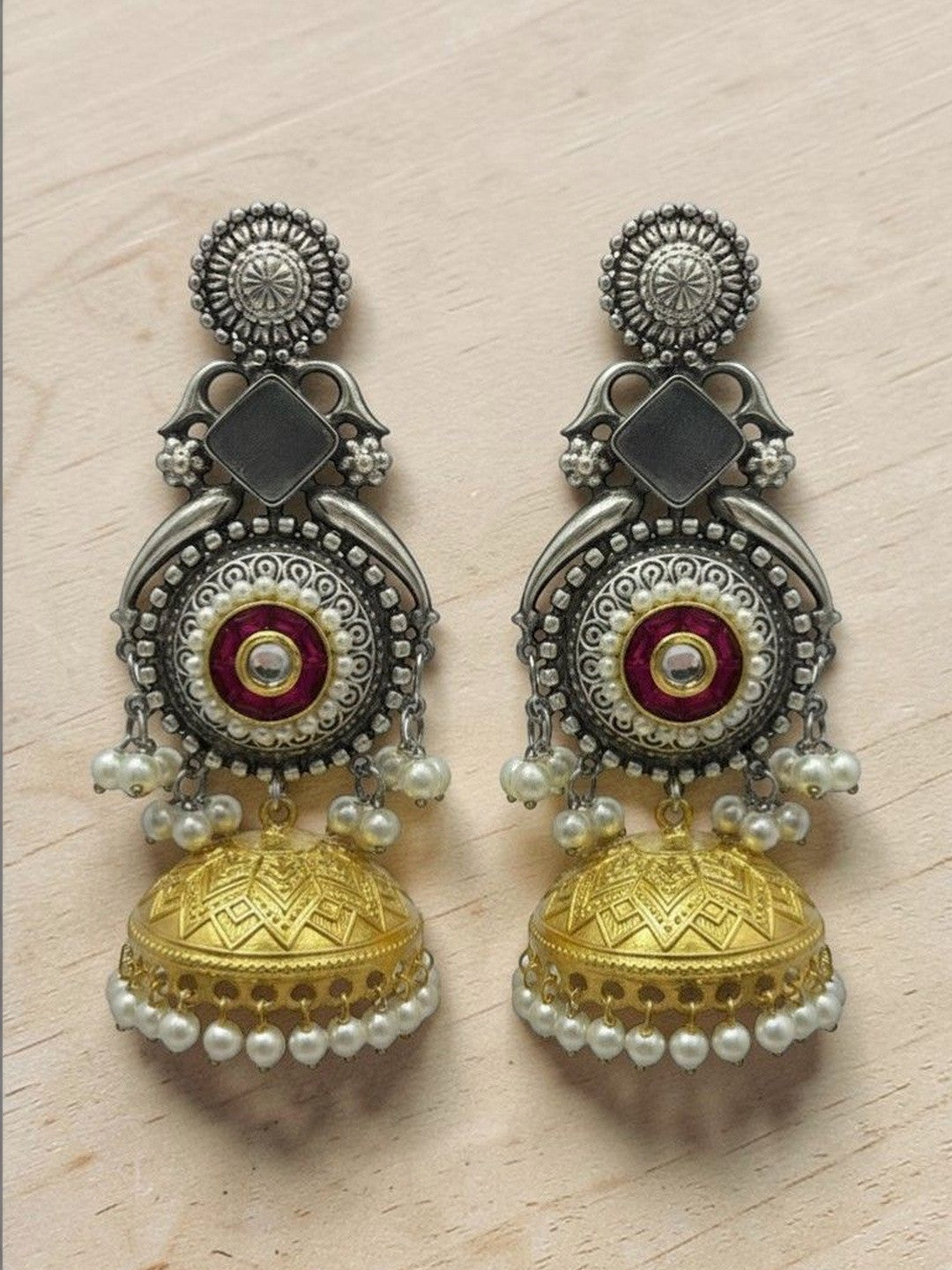 Oxidised silver and gold-tone jhumka earrings with red enamel kundan centre displayed on a wooden surface