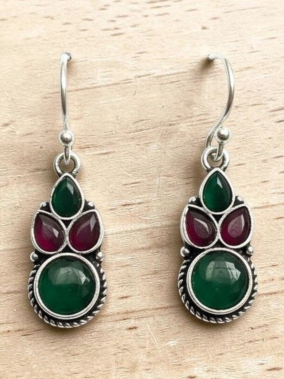 Pair of oxidised silver drop earrings with green and ruby-red pear and round stones on a wooden surface