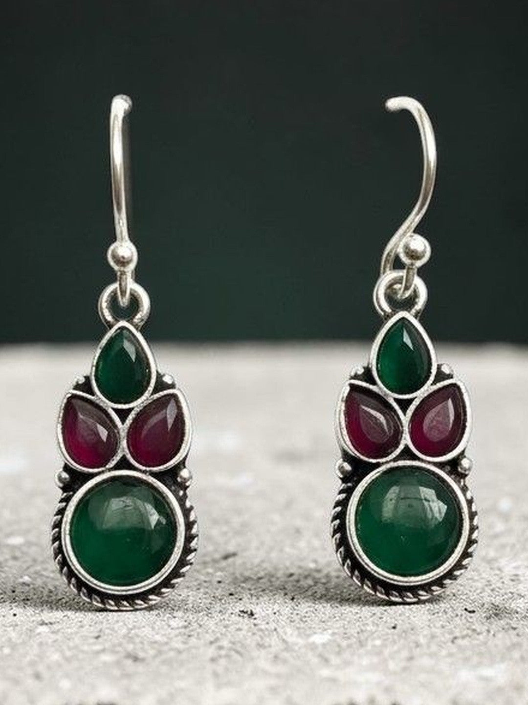 Close-up of oxidised silver green and red stone drop earrings against a dark teal background