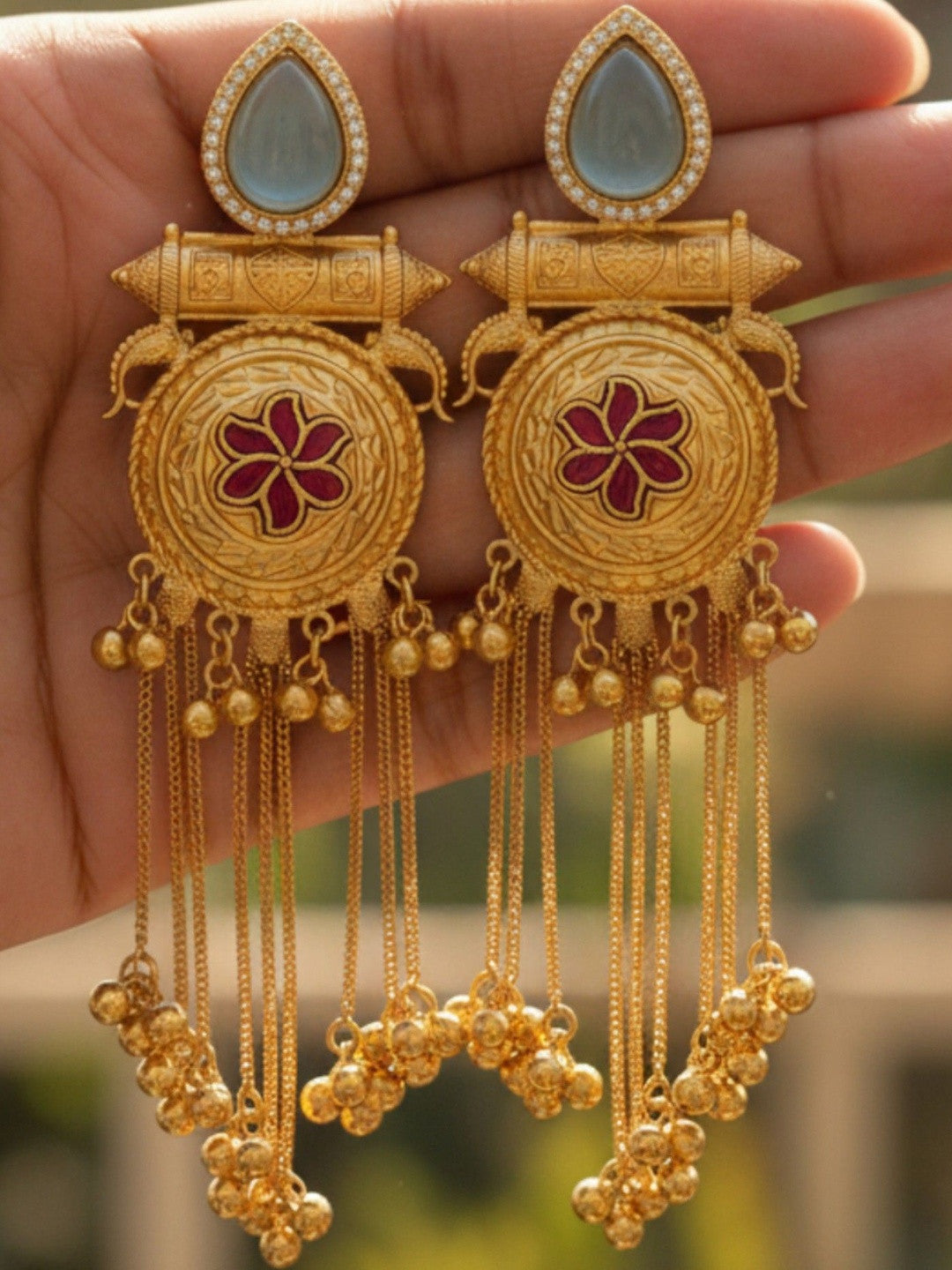 Pair of gold-plated chandbali earrings held in palm displaying blue teardrop stones and layered chain tassels