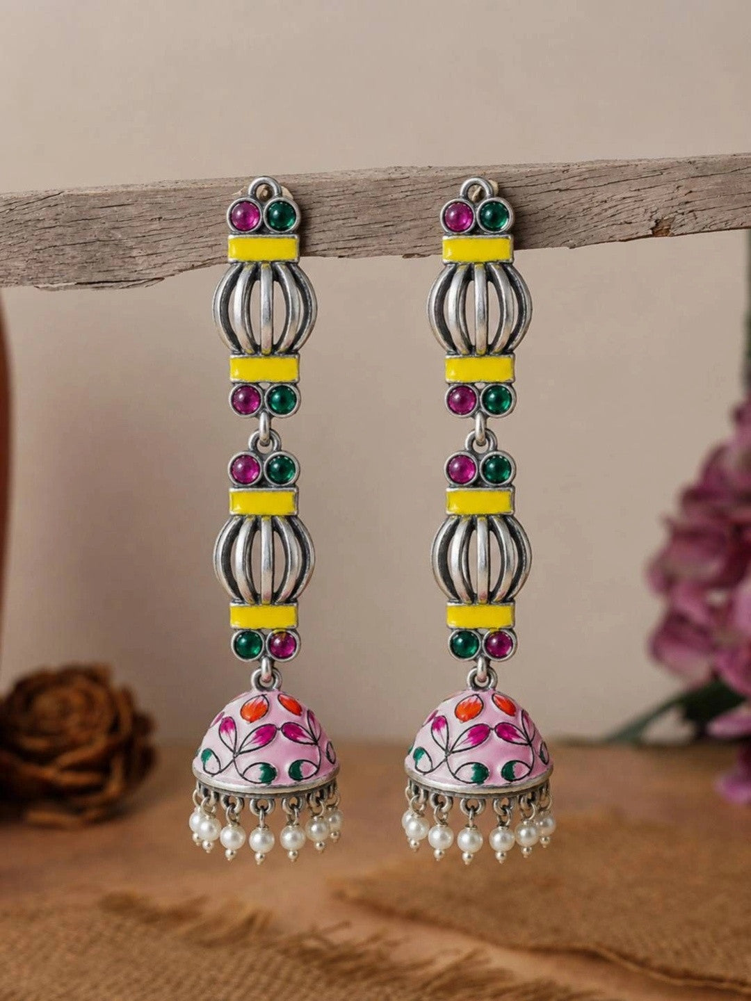 Pair of oxidised silver jhumka earrings with yellow enamel bars and pink floral dome, hanging on a wooden display rod