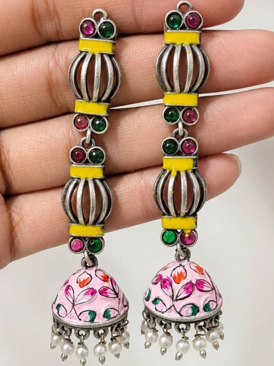 Close-up of jhumka earring pair resting on a palm showing hand-painted floral enamel dome and pearl bead fringe