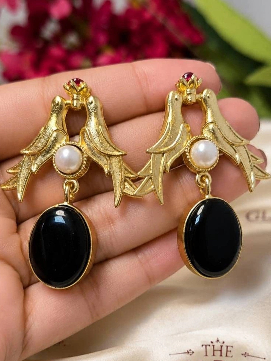 Close-up of gold swallow bird earrings with black oval drop and red stone crown detail held in palm