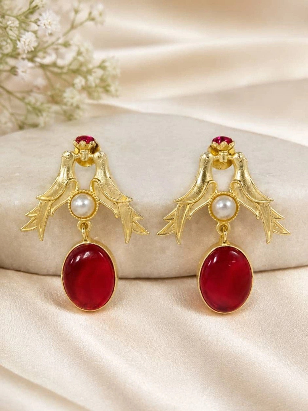 Pair of gold-plated love birds drop earrings with red oval stone and pearl on a smooth stone surface