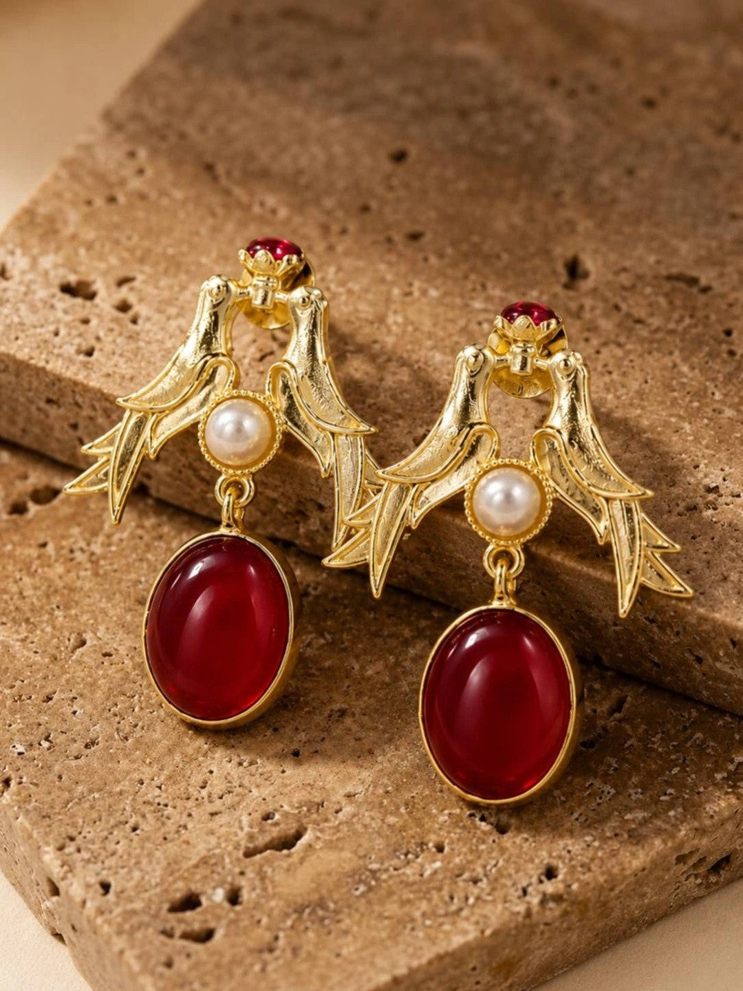 Close-up of gold love birds earrings with faux pearl centre and deep-red drop stone on textured travertine