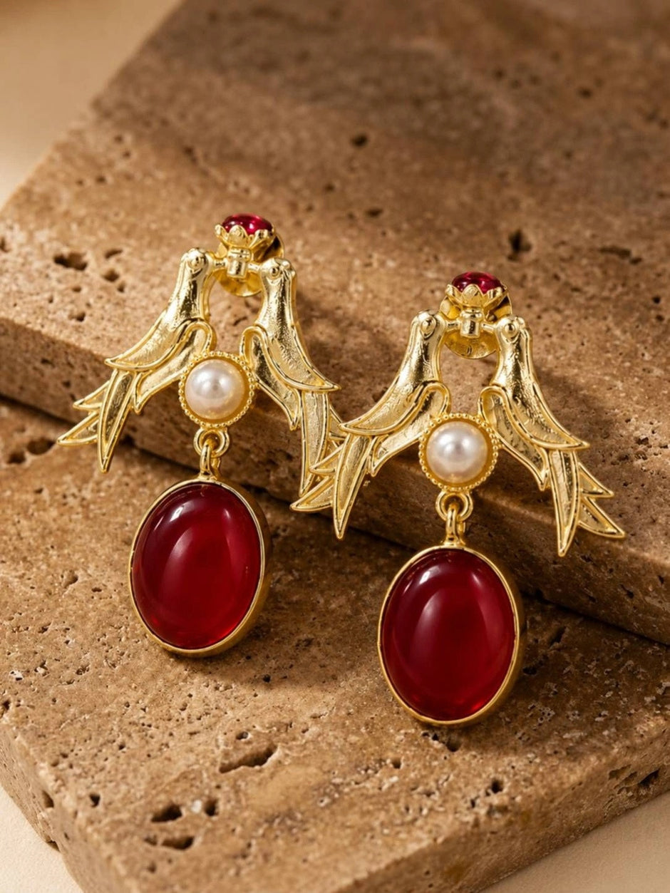 Close-up of gold love birds earrings with faux pearl centre and deep-red drop stone on textured travertine