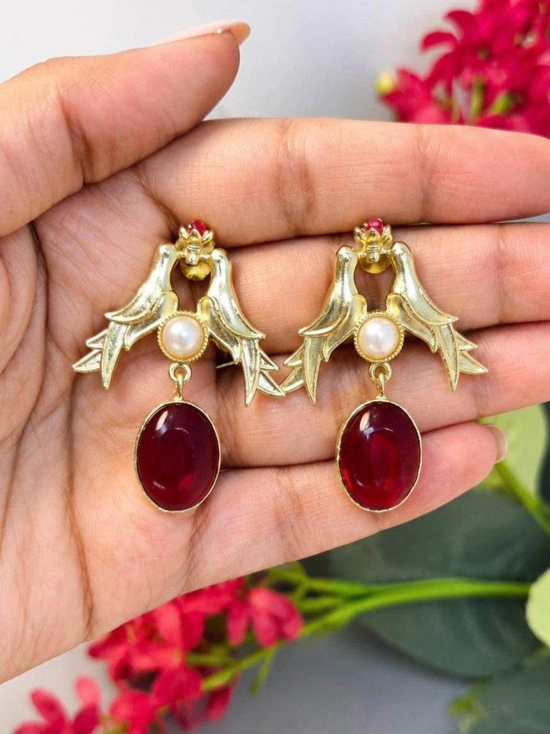 Gold love birds drop earrings held in an open palm showing full scale and red oval stone detail