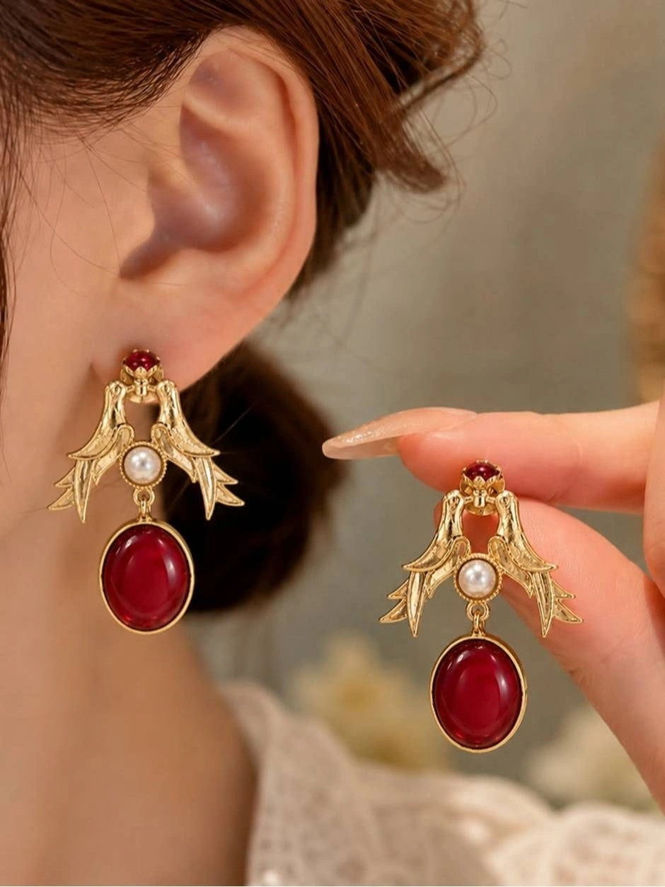 Gold-plated bird drop earring worn on ear alongside second earring held between fingers showing red stone