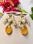 Gold-plated love birds earrings with honey amber oval drops displayed on beige jewellery cushion