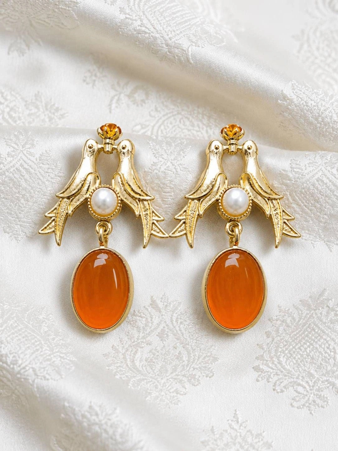 Pair of gold-plated love birds drop earrings with pearl centre and oval orange resin drop on white fabric