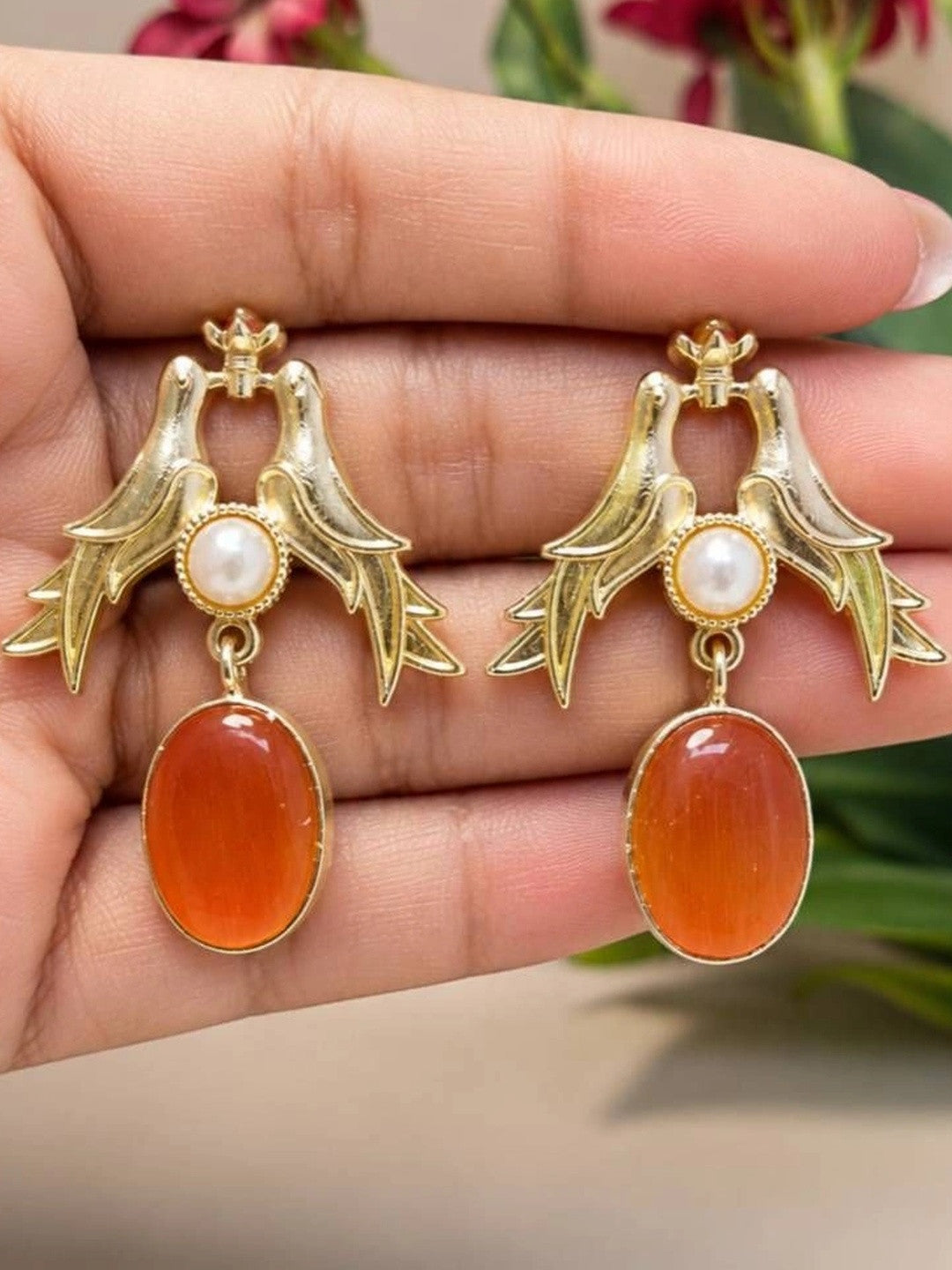 Close-up of two gold love birds earrings with pearl and carnelian resin drops resting on a hand