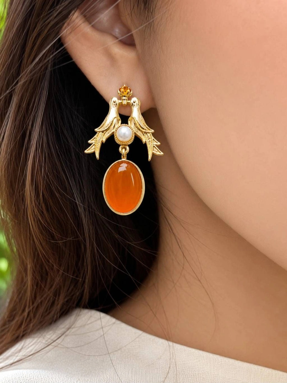 Woman wearing one gold love birds earring with orange oval drop against her ear, side profile view