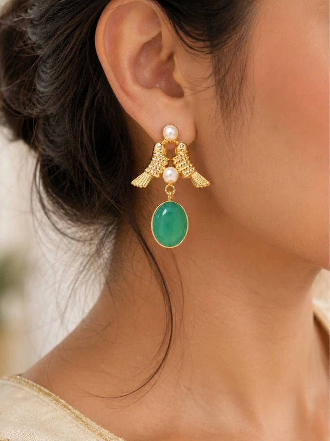 Gold-plated twin bird earring with green stone drop worn on a woman's ear against a soft blurred background