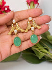 Two gold-plated twin bird earrings with green oval stone drops resting on an open palm with green leaves behind