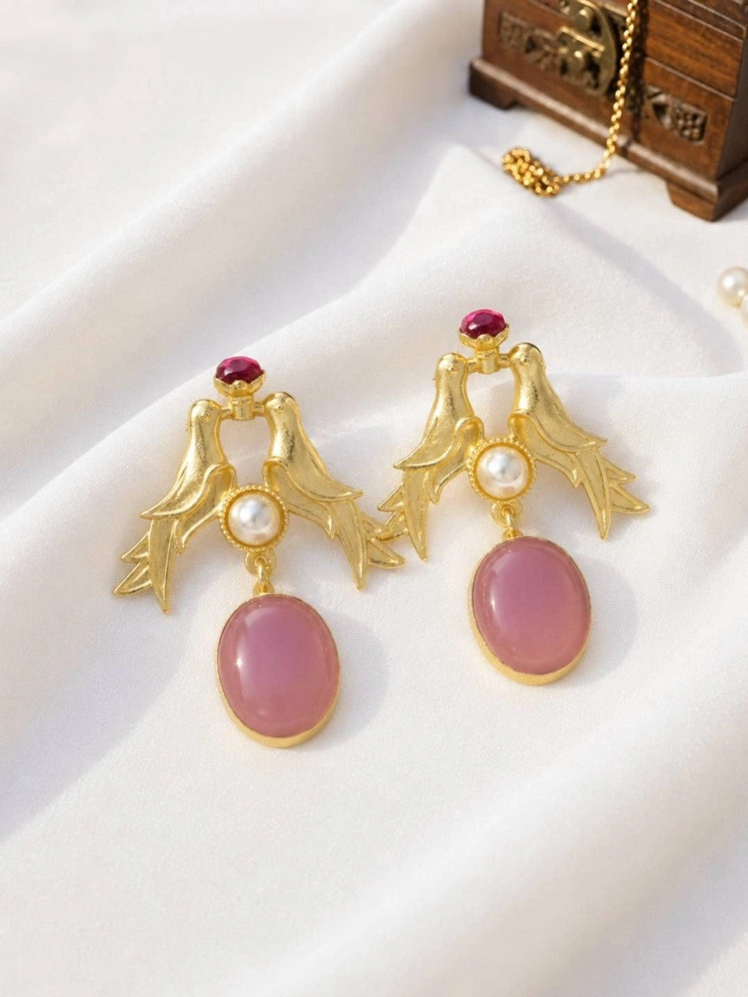 Pair of gold-plated love bird drop earrings with pink oval cabochon and pearl accent on white fabric