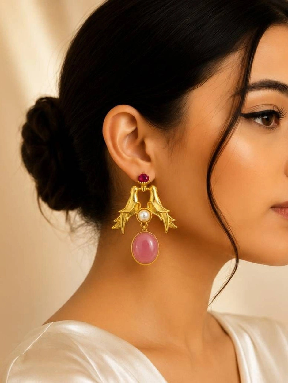 Woman wearing gold love bird drop earring with pink oval stone against a beige background