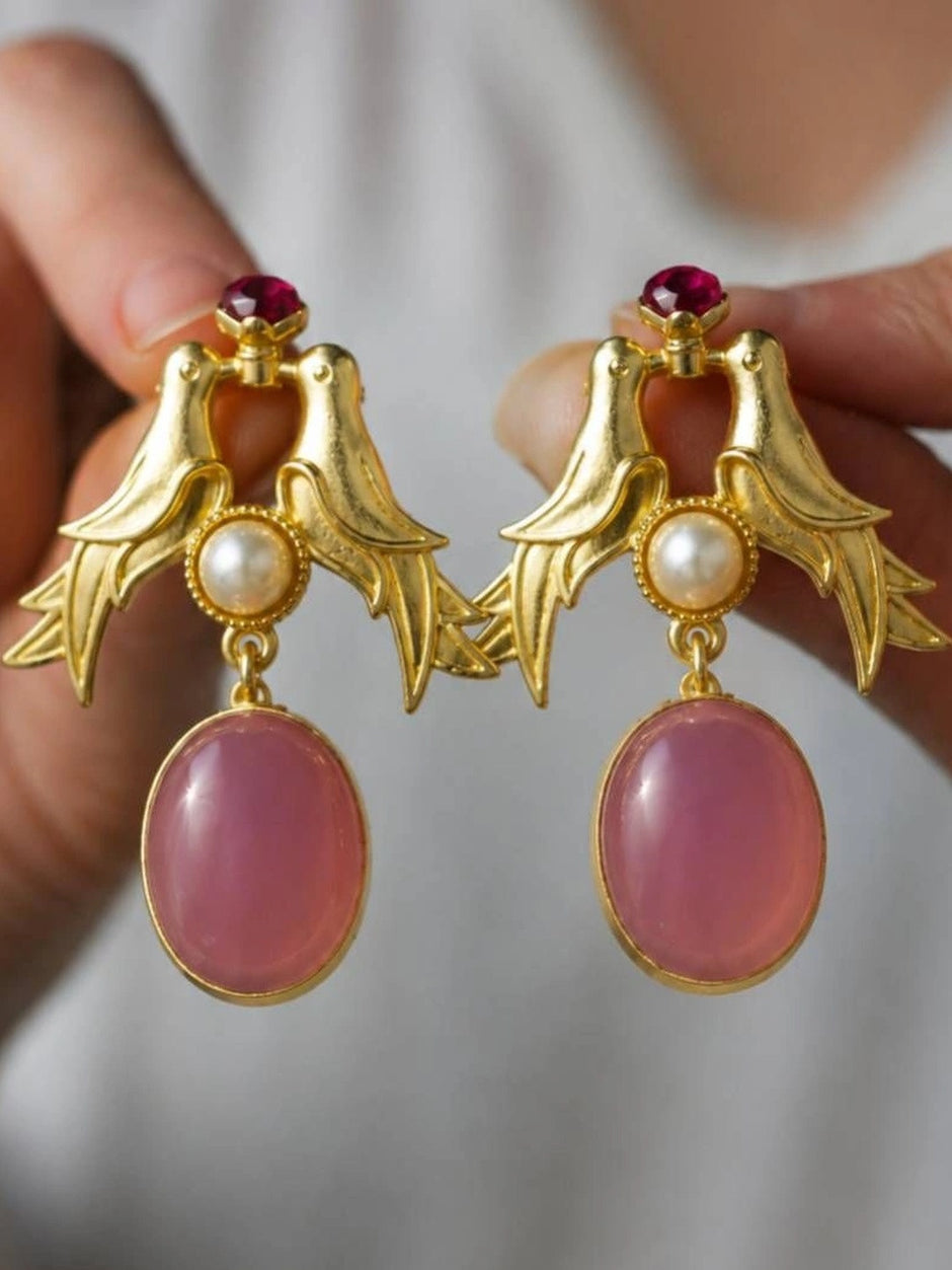 Hand holding a pair of gold-plated love bird earrings showing pink cabochon drops and red stud tops