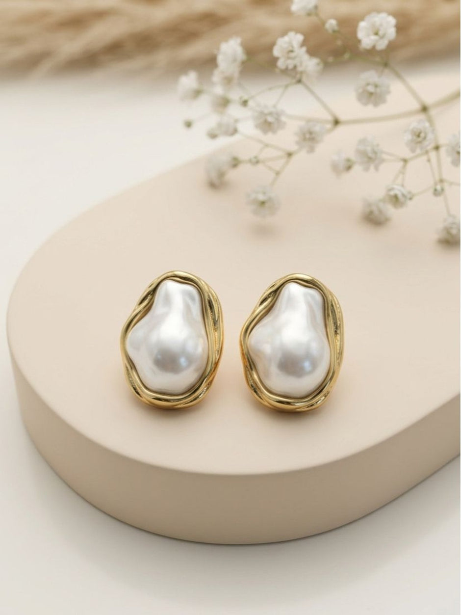 Two gold-plated baroque pearl studs displayed on a beige ceramic stand with dried flowers