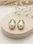 Two gold-plated baroque pearl studs displayed on a beige ceramic stand with dried flowers