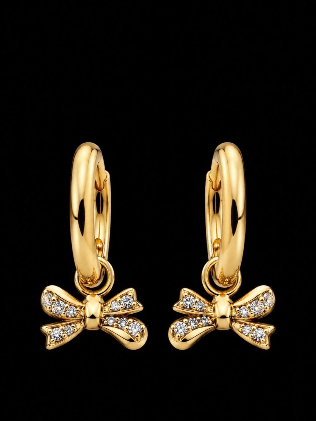 Pair of gold-plated huggie hoop earrings with pavé cubic zirconia bow charms on black background